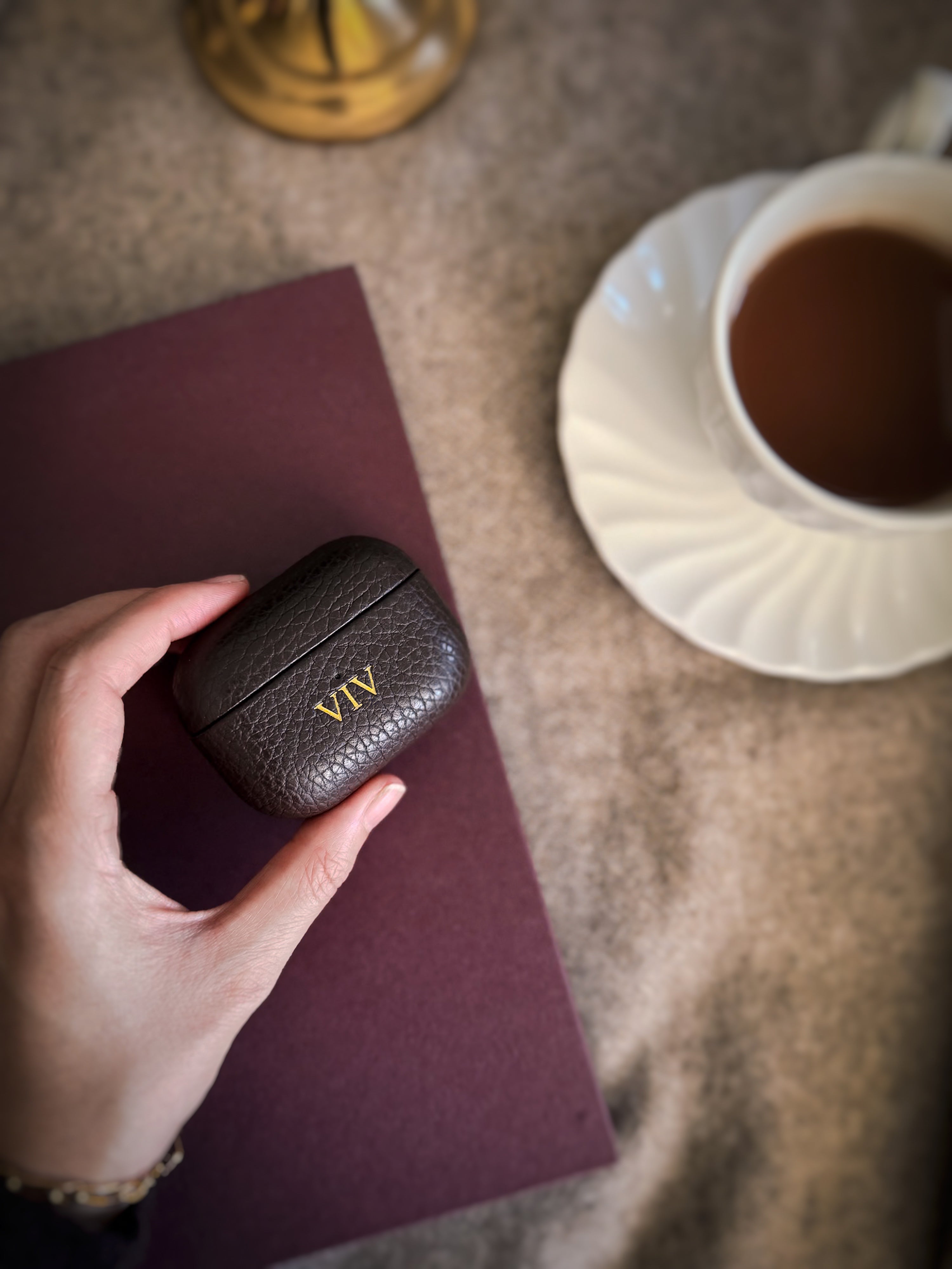 Person holding a textured black leather AirPod case with 'VIV' initials, next to a cup of tea on a saucer.