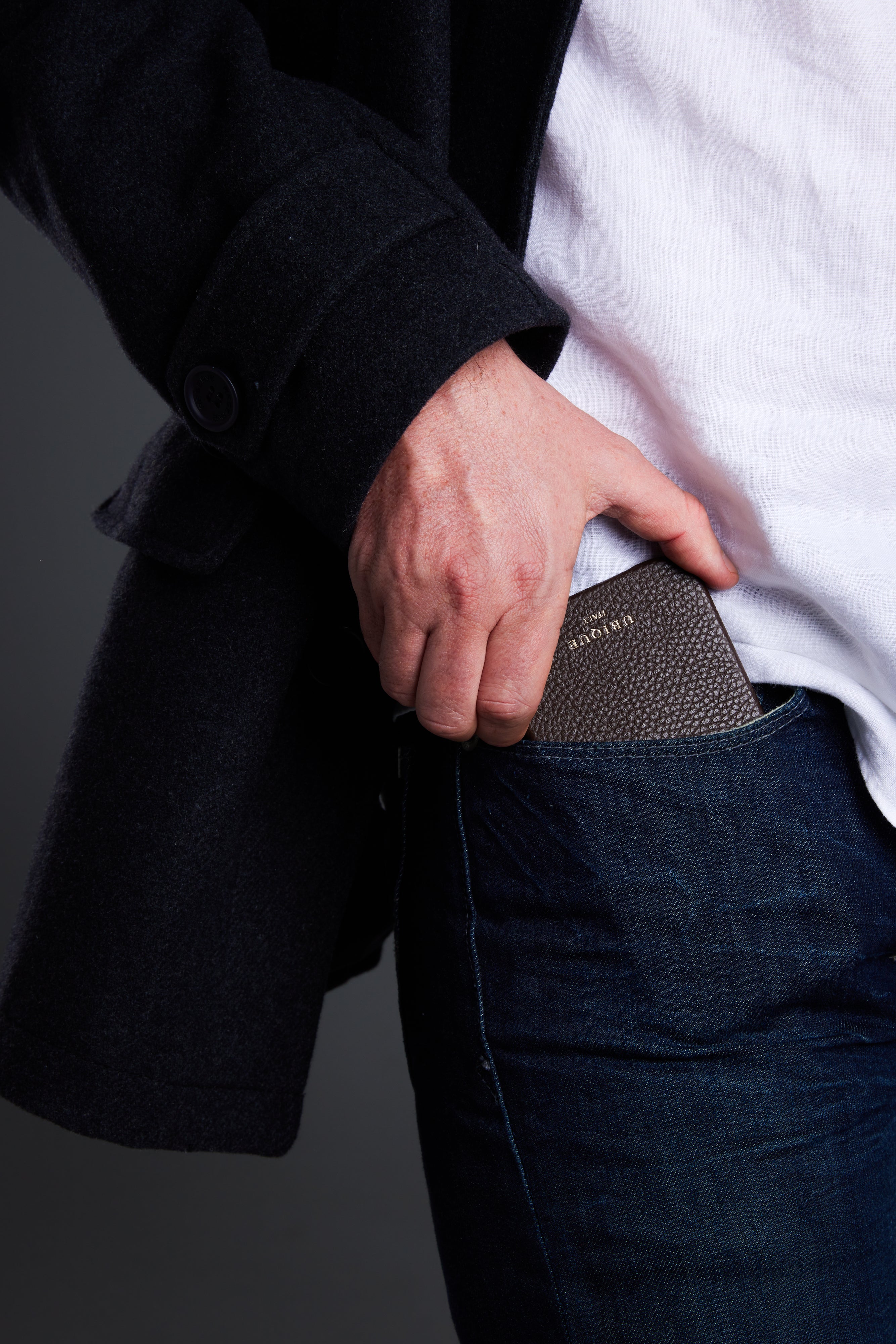 Person wearing a black coat and white shirt, with a pebble grain leather iPhone case in their pocket against a gray background.