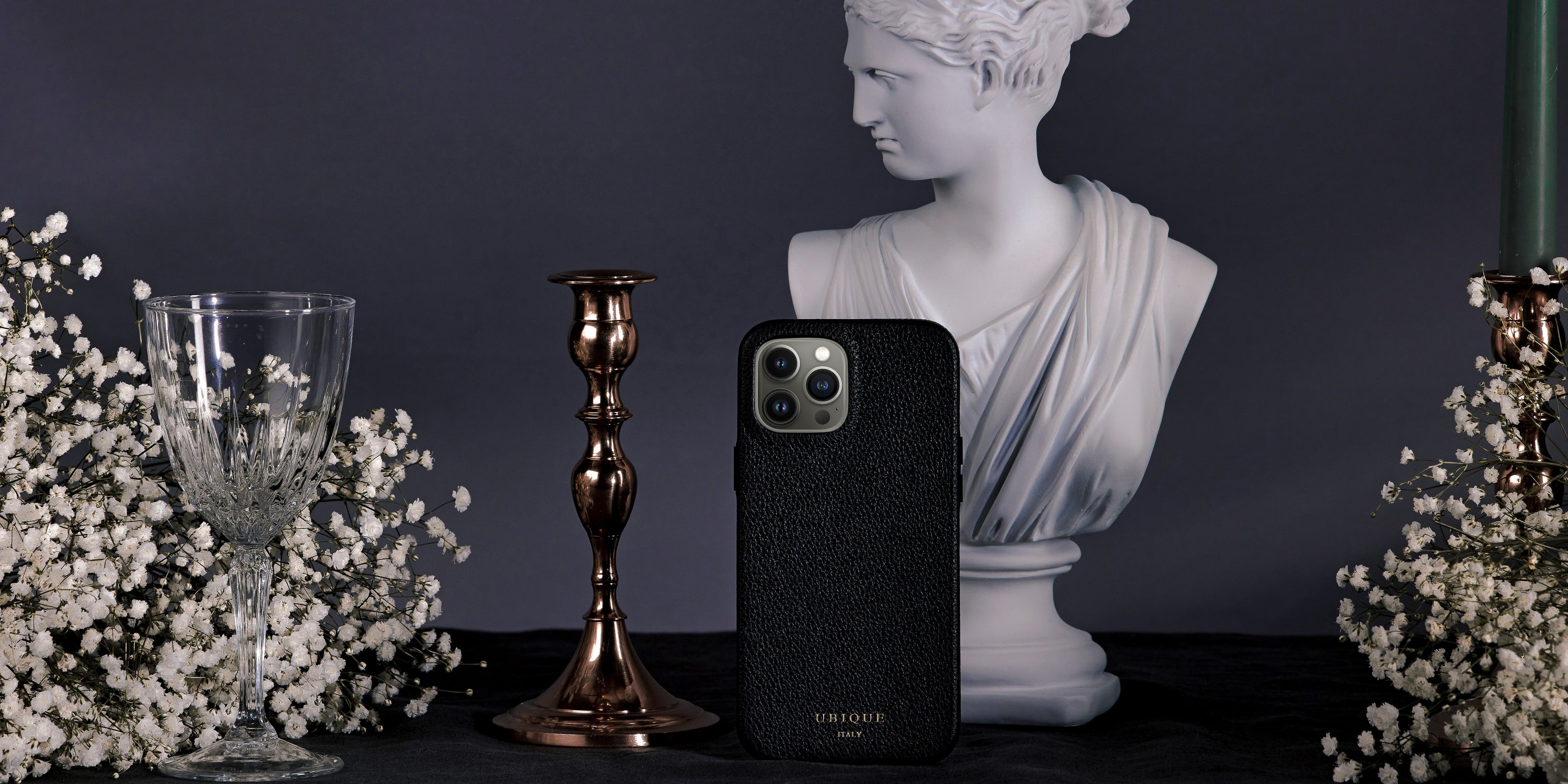 Black leather iPhone case with white statue bust and rose gold candle holder with white flowers on a dark fabric