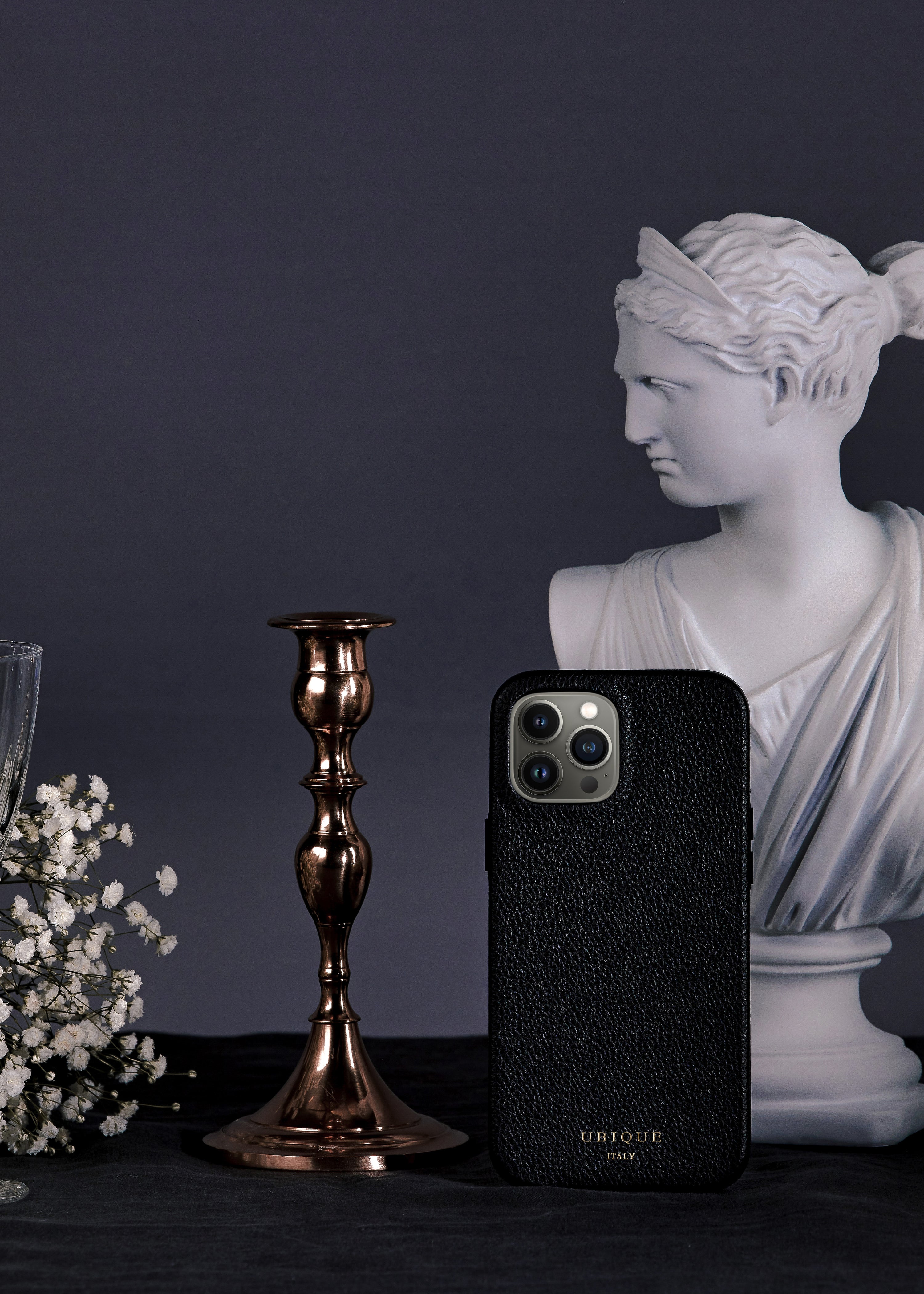 Black leather iPhone case with white statue bust and rose gold candle holder with white flowers on a dark fabric